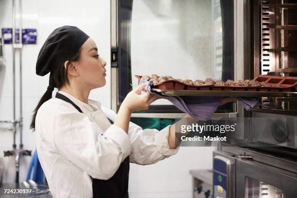 side view of female chef taking out tray of cookies from oven at commercial kitchen - chef cooking stockfoto's en -beelden