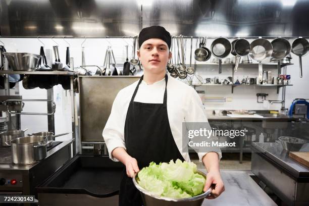 portrait of confident male chef holding container of vegetables at commercial kitchen - kochlehrling stock-fotos und bilder