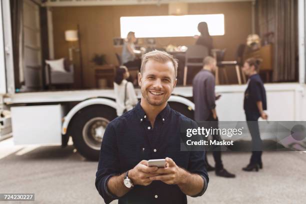 portrait of smiling businessman holding mobile phone with colleagues and portable office truck on road in background - mid aged stock pictures, royalty-free photos & images