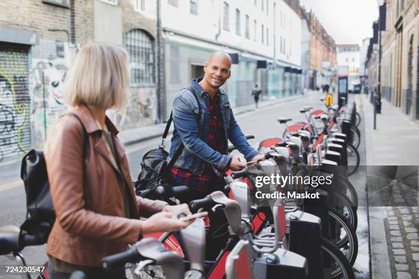 multi-ethnic couple renting bicycles from bike share stand in city - bicycle rental stock pictures, royalty-free photos & images