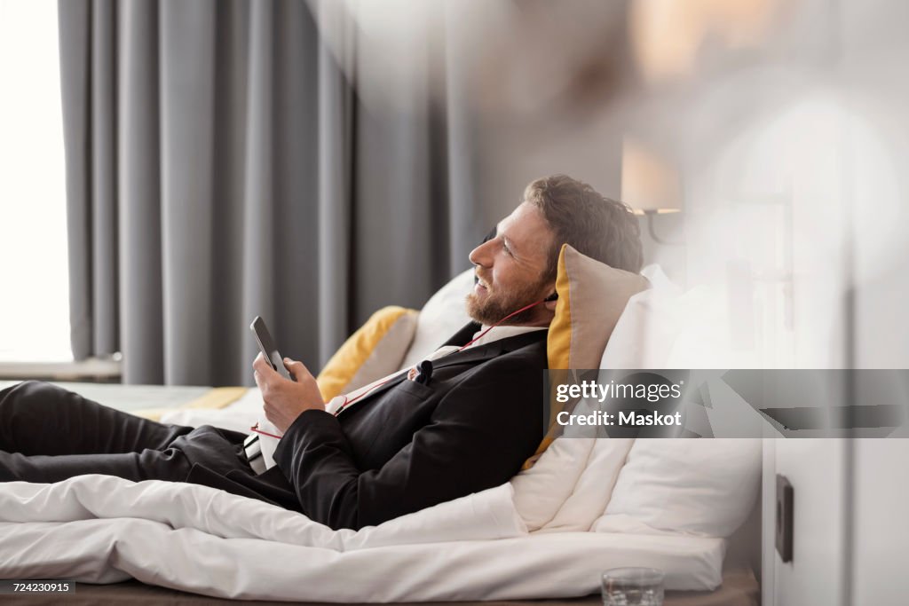 Side view of businessman listening to music while lying on bed at hotel room