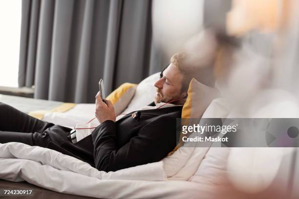 side view of businessman listening music while lying on bed at hotel room - bed side view stock pictures, royalty-free photos & images