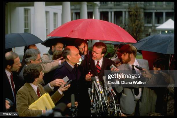 Senator Don Riegle with Senator Jake Garn, sheltered under big red umbrella, talking to reporters.