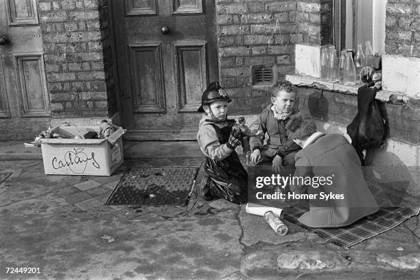 Three young boys playing on a tartan travel rug in a street in Elephant and Castle, London, 1970. They are on their family doorstep with plastic...