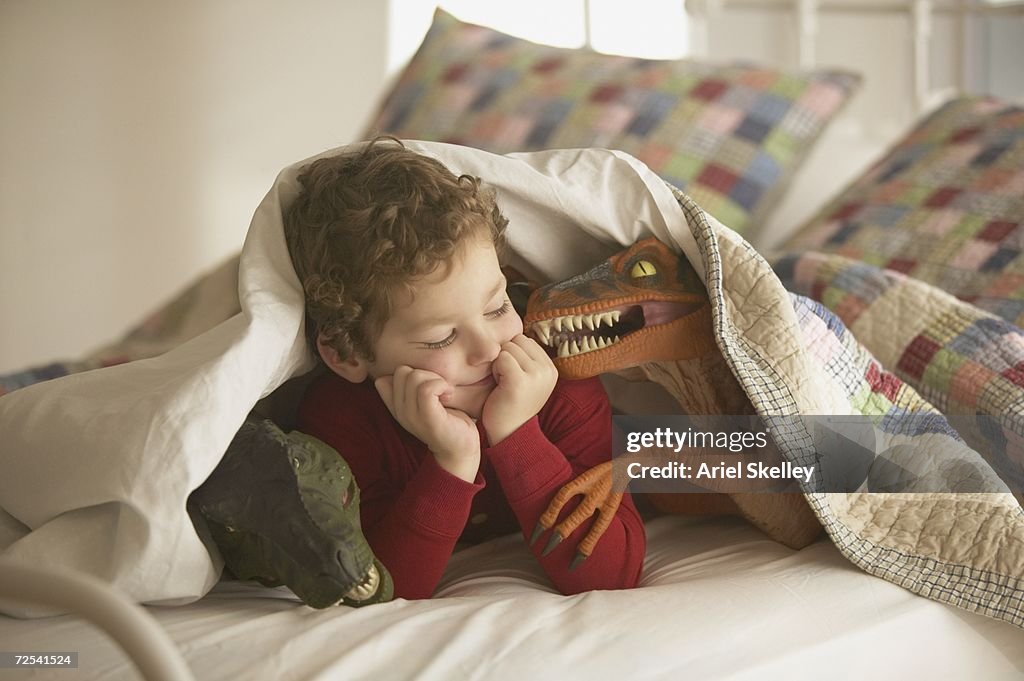 Young boy under covers in bed with toy dinosaurs