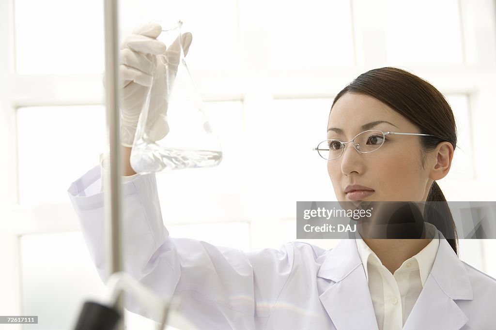Female scientist holding up and watching a conical flask, front view, differential focus