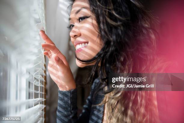 mixed race woman peeking out window behind blinds - peep window stock pictures, royalty-free photos & images