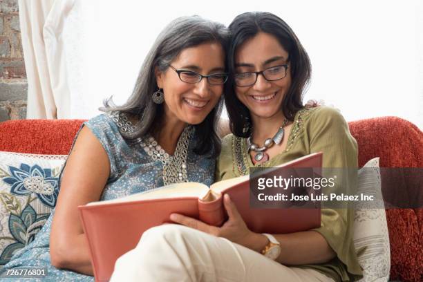 smiling mother and daughter sitting on sofa reading book - indian mother daughter stock-fotos und bilder