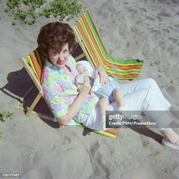 caucasian mother sitting on beach feeding bottle to baby son - 1960s baby stockfoto's en -beelden