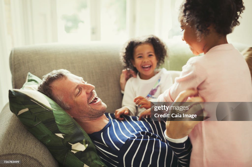 Playful multi-ethnic father and daughters tickling and laughing on sofa