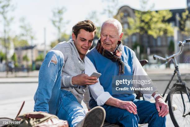 senior man and adult grandson on a bench looking at cell phone - alter mann jung geblieben stock-fotos und bilder