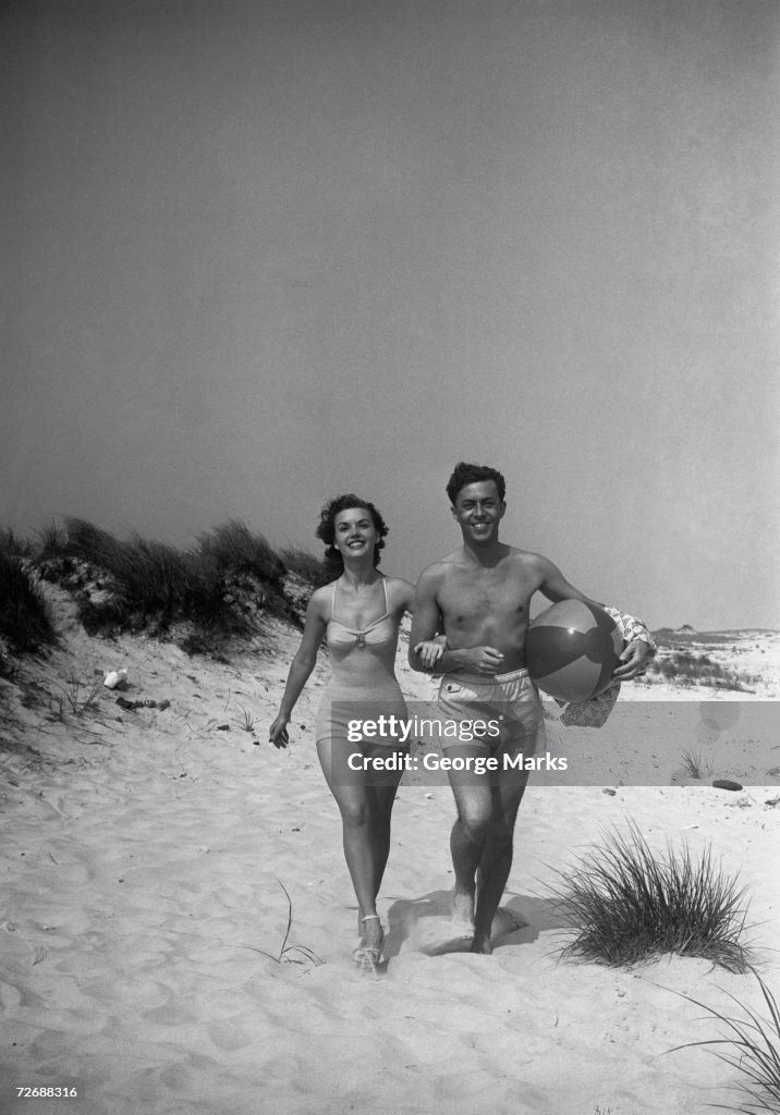 Couple walking on beach, man carrying ball, (B&W)