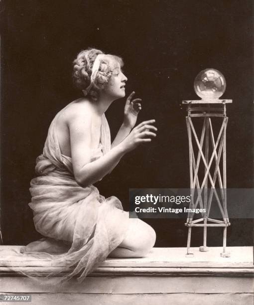 Young woman wrapped in a diaphanous garment gestures as she kneels before a small table on which sits a crystal ball, early 20th Century.