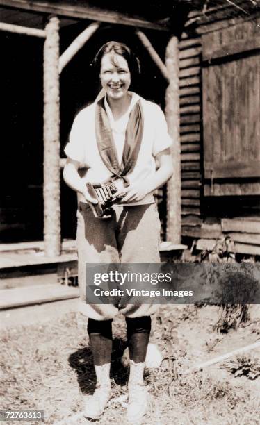 Woman smiles and holds a folding pocket camera outside of a wooden structure, 1920s.