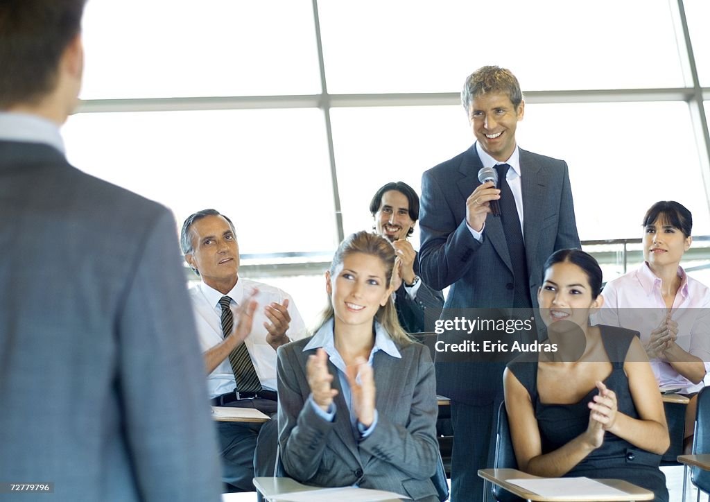 Executives in seminar, one man holding microphone, others clapping