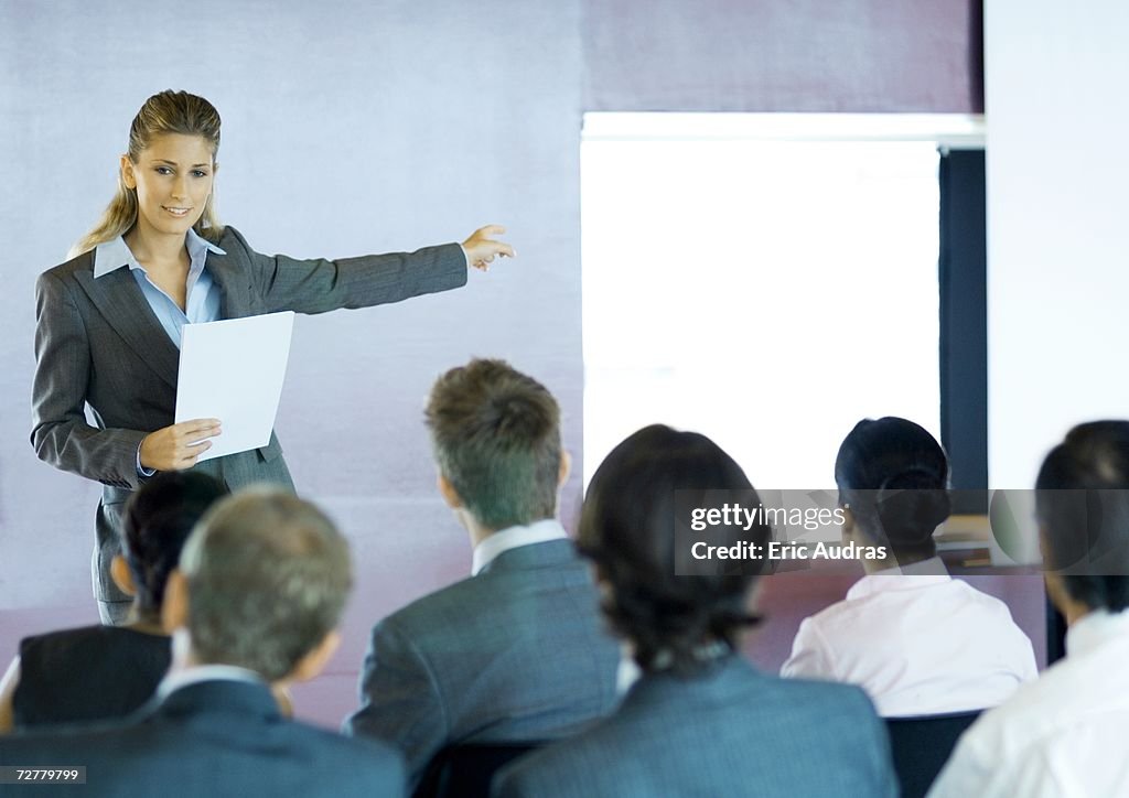 Executives sitting in seminar, woman standing facing group, gesturing to screen