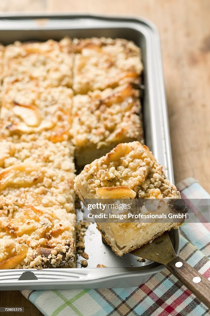 Apple crumble cake in baking tin with piece on server