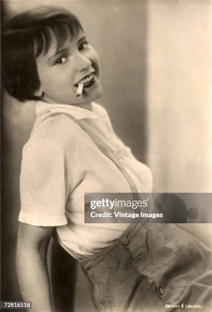 American actress Louise Brooks poses in lederhosen and smokes a cigarette in this postcard image from von Zander & Labisch, Germany, late 1920s or...