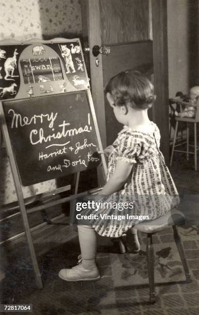 Little girl uses a piece of chalk to add her final touches to a Christmas message on a small blackboard, early 20th Century. The message is written...