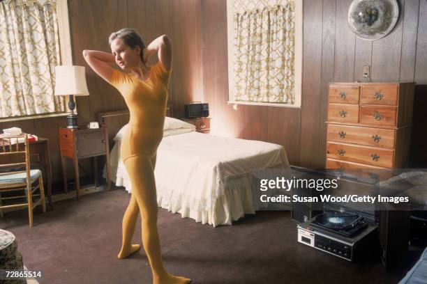 An unidentified young woman, dressed in a yellow leotard and matching tights, exercises in her wood-panelled room, to the music from a record player...