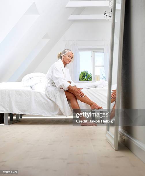 mature woman sitting on bed in bathrobe, putting cream on her leg - legs stockfoto's en -beelden