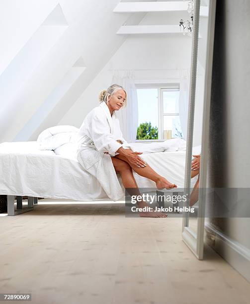 mature woman sitting on bed in bathrobe, putting cream on her leg - leg stock pictures, royalty-free photos & images