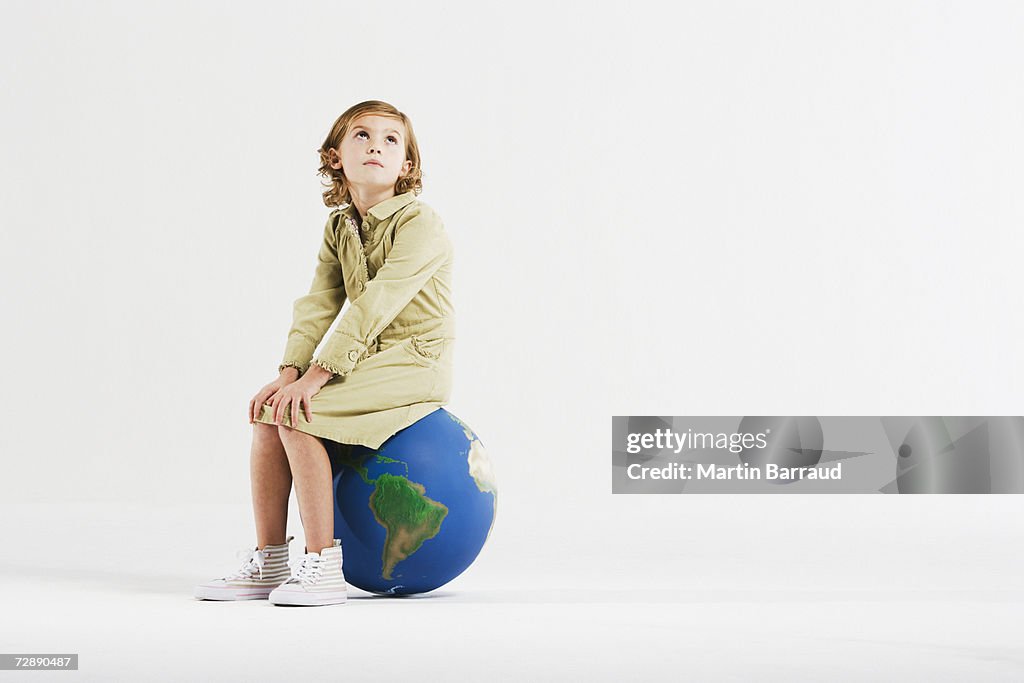 Girl (8-9) sitting on globe in empty white room, looking upwards