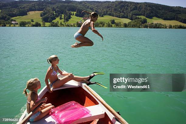 girls (8-15) sitting on boat, one jumping in water, side view - northern european descent stockfoto's en -beelden