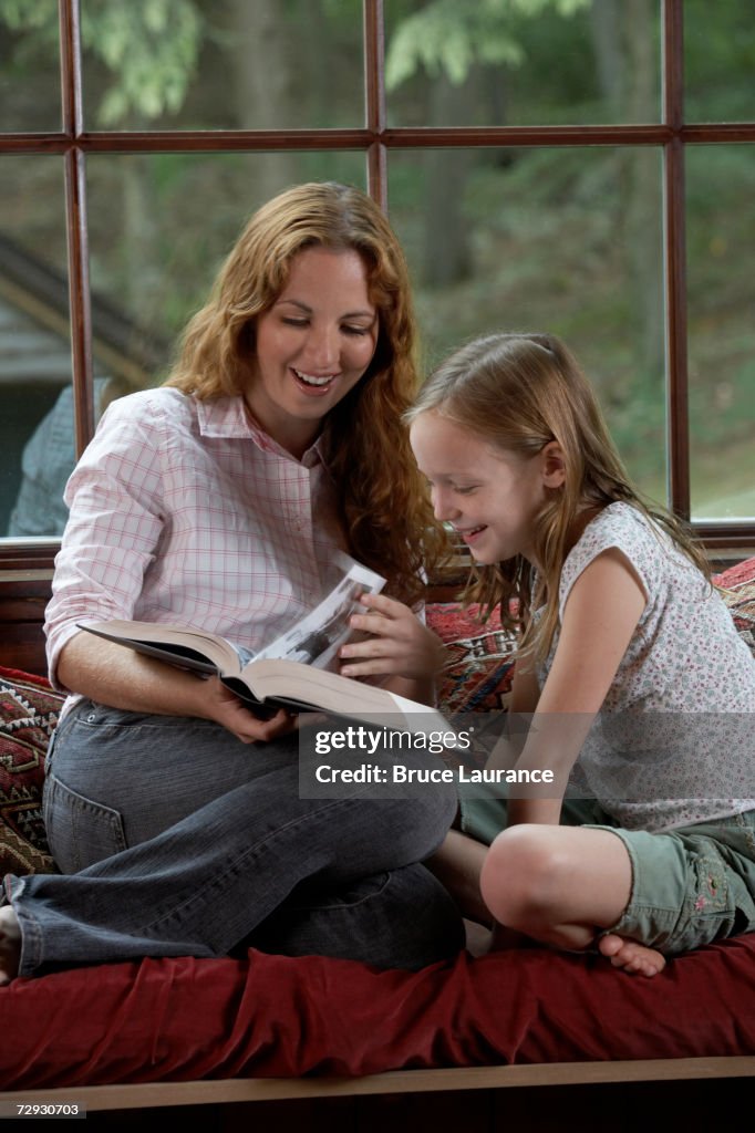 Mother and daughter (8-9) sitting on sofa, reading book, smiling