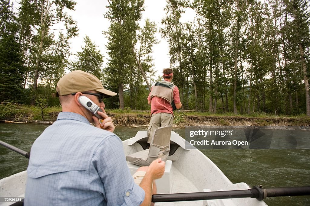 Young man using cell phone while fly-fishing on the Elk River from a dory, Fernie, East Kootenays, British Columbia, Canada.