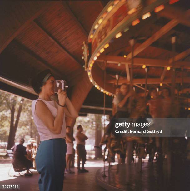 An unidentified woman smiles as she films children on a carousel with a portable camera, 1950s.