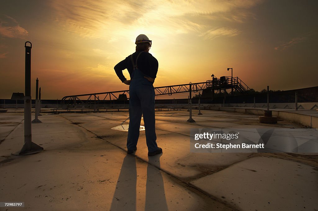 Man in safety clothing standing on top of oil tank at sunrise rear view