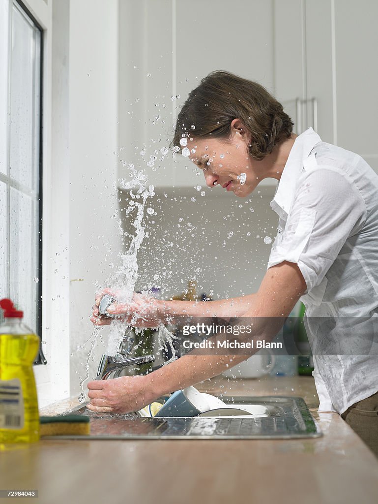 Woman at sink in domestic kitchen being drenched by broken tap, side view