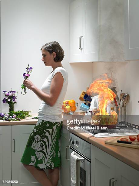 "woman leaning against kitchen worktop holding flower, frying pan on fire behind" - ignorance foto e immagini stock