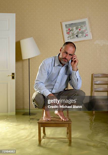 mature man using mobile phone crouching on chair in flooded living room - flooded home stock pictures, royalty-free photos & images