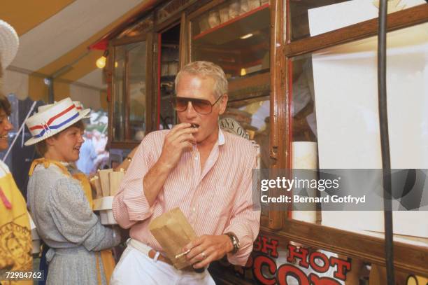 Portrait of American movie actior and philanthropist Paul Newman as he eats from a bag of popcorn and leans against a popcorn stand that stocks his...