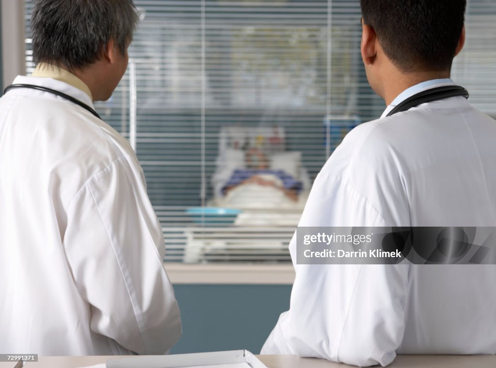 Two male doctors looking at patient lying on bed in ward, rear view