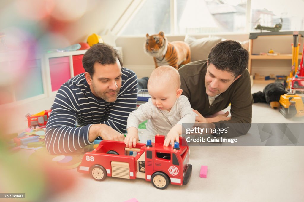 Male gay parents and baby son playing with fire engine toy in playroom