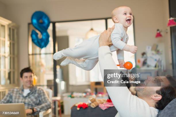 Male gay parents lifting and playing with baby son in living room