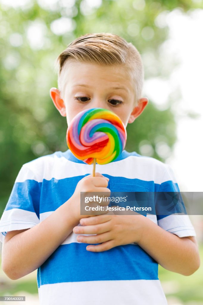 Boy (6-7) squinting and holding big, colorful lollipop
