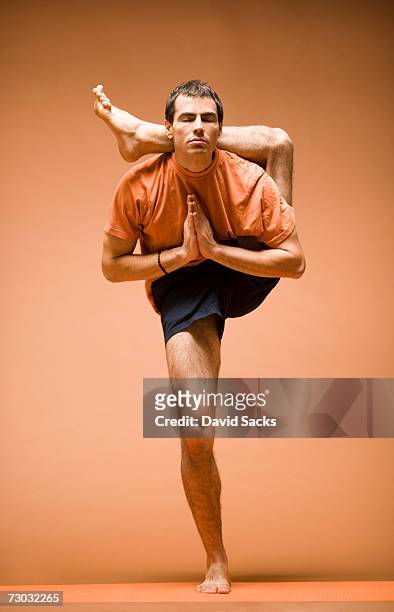 young man standing with leg behind head in yoga pose, front view - yoga stock-fotos und bilder
