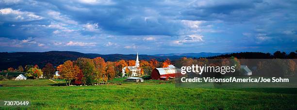 this is a view of the village in autumn. there is a typical new england white church with a tall steeple and a red barn. there is a vast green field in front of the view of the village with fall foliage in the village. - peacham stock pictures, royalty-free photos & images