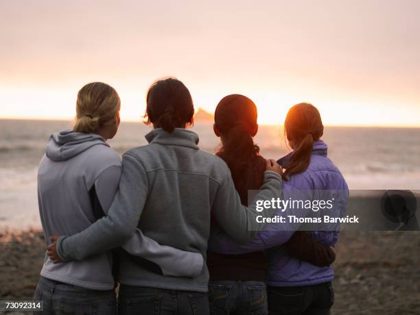 four young women, arms around each other, looking out to sea, rear view - arms around each other foto e immagini stock