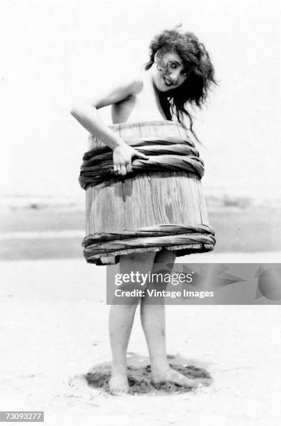 Young woman, one of Mack Sennett's 'Bathing Beauties,' covers her nakedness with half a barrel, on a beach, 1910s.