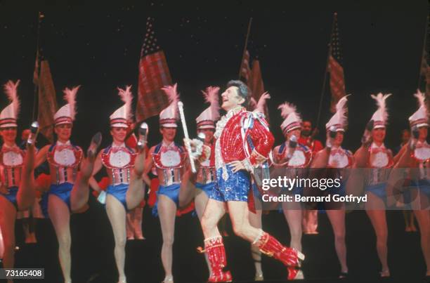 American musician and performer Liberace prances onto the stage in sequined short shorts in front of a kick line of the Rockettes, Radio City Music...