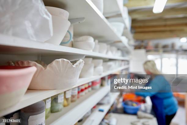 a woman working at a bench in a pottery studio. shelves storing pots and bowls. - avondschool stockfoto's en -beelden