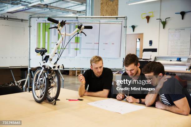 three men in a meeting at a bicycle factory, sitting at a table with a folding bicycle on the tabletop. - klapprad business stock-fotos und bilder