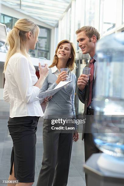 three colleagues talking by water cooler in office - dispensador de agua fotografías e imágenes de stock