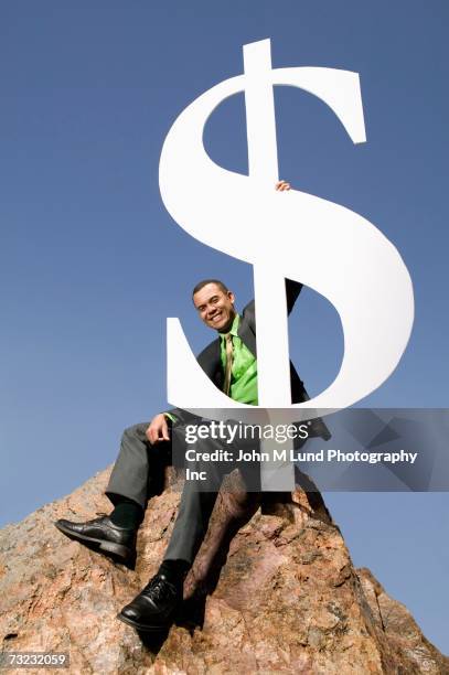 businessman sitting on boulder holding large dollar sign - impact summit stock pictures, royalty-free photos & images