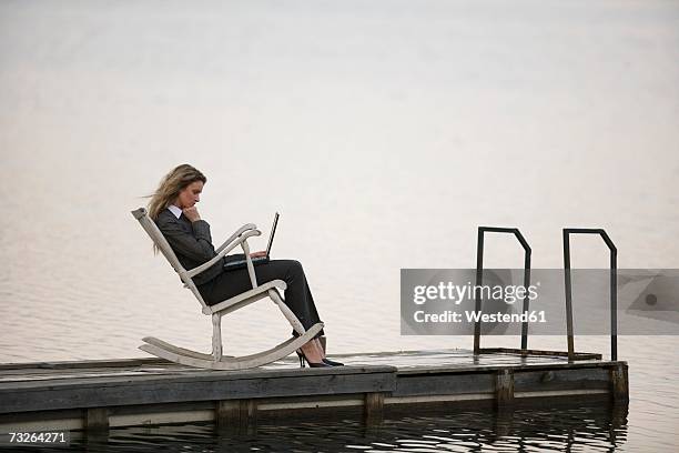 businesswoman sitting in rocking chair on jetty, using laptop, side view - schaukelstuhl stock-fotos und bilder